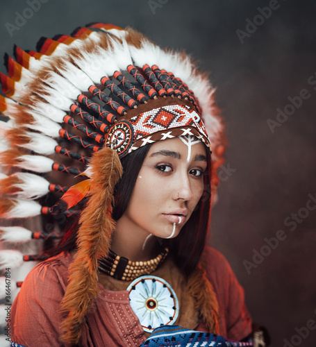 Plakat Close up portrait of a attractive young American Indian woman with make up in traditional feather hat and clothes on a dark background