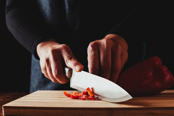 Hands cut red pepper on a wooden board.  The cook uses an apron.  Photo on black background