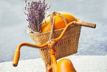 Knitted Straw Basket With Cheese Head And Bouquet Of Dry Lavender Inside, Attached To Handlebars Of An Orange Retro Bicycle On Light Background. Point Of View Shot. Concept Of Natural Eco Materials.