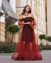 Elegant Fashionable Female Model In Red Long Evening Dress With Open Shoulders And Sleeves Standing And Posing At City Street. Beautiful Caucaisan Woman With Wavy Hair And Makeup Outdoors Portrait