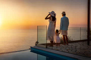 Eine elegante Familie im Urlaub steht zusammen am Pool und genießt den idyllischen Sonnenuntergang über dem Meer