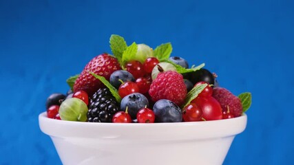 Wall Mural - Mix of wild forest berries in bowl on blue background, collection of strawberry, blueberry, raspberry and blackberry, 4K UHD video footage