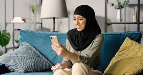 Wall Mural - Portrait of beautiful sick arab woman in hijab at home using smartphone while sitting on sofa.