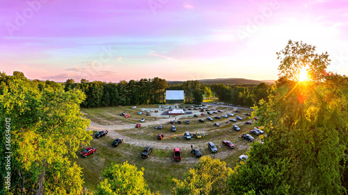 Outdoor Drive-In Movie Theater with Pink Sunset. Aerial drone view looking over cars from back of parking lot towards white movie screen.