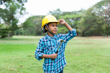 Close-up of young boy wearing engineer clothes and holding steel hammer, wearing yellow safety helmet standing in the garden while field trip or summer camp.