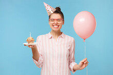 Young Happy Birthday Girl In Hat, Holding Pink Balloon In One Hand And Holiday Cupcake In Another, Smiling With Closed Eyes, Isolated On Blue Background