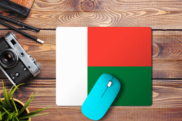Madagascar flag on wooden background with blue wireless mouse on a mouse pad, top view. Digital media concept.