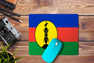 Wall Mural - New Caledonia flag on wooden background with blue wireless mouse on a mouse pad, top view. Digital media concept.