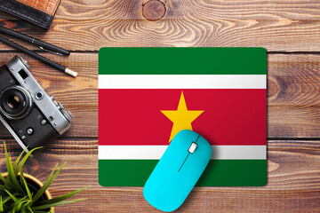 Wall Mural - Suriname flag on wooden background with blue wireless mouse on a mouse pad, top view. Digital media concept.