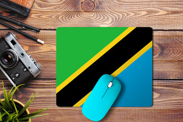 Wall Mural - Tanzania flag on wooden background with blue wireless mouse on a mouse pad, top view. Digital media concept.