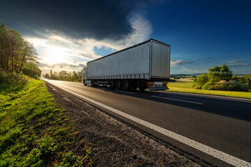 White truck driving on the asphalt road in rural landscape at sunset with dark storm cloud