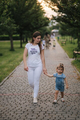 Wall Mural - Beautiful young mom walkin in park with her adorable little daughter. Cute little girl hom mothers's hand