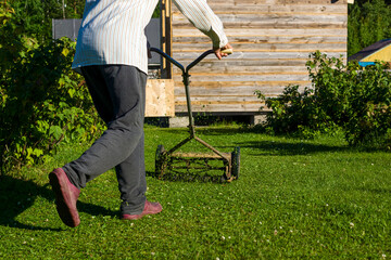 Country man mows grass at a farm with retro mechanical lawn mower in evening