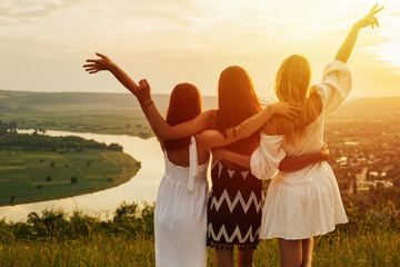 Wall Mural - The back of three  girls hugging together on the mountain. Happiness and togetherness concept. Beautiful summer evening. View from the back. Concept of freedom, rest and relaxation.