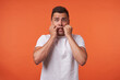 © timtimphoto - Studio photo of young pretty brown haired male holding raised hands on his cheeks while looking scaredly at camera, standing against orange background