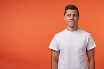 Afraid young short haired brunette guy dressed in casual wear biting worringly lips while looking aside, keeping hands along body while posing over orange background