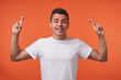 © timtimphoto - Good looking young attractive brown haired man in white t-shirt smiling with closed eyes and raising hands with crossed fingers, posing over orange background