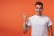 © timtimphoto - Joyful young handsome short haired brunette male looking happily at camera with wide smile while thumbing aside with raised hand, isolated against orange background