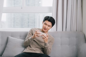 Wall Mural - Asian young man is seriously playing the game on his smartphone on the couch.