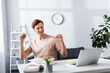 © LIGHTFIELD STUDIOS - Positive freelancer showing yeah gesture near laptop and stationery on table at home, earning online concept