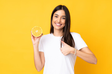 Wall Mural - Young caucasian woman holding an orange isolated on yellow background with thumbs up because something good has happened