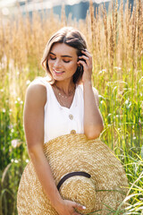 Wall Mural - Young smiling woman holding her big straw hat and looking down while standing on the field