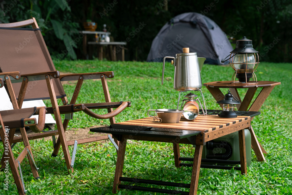 Stainless steel kettle, portable gas stove, bowl and vintage lanterns with outdoors table set on green lawn in camping area - obrazy, fototapety, plakaty 