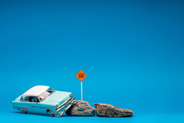 Light blue, retro car figurine next to two boulders and a construction zone sign, on blue background.