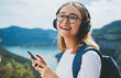 portrait smiling traveler woman with glasses holds smartphone device in hands and listens music for headphones via Internet technology to enjoy hiking walk  on top of mountains during freedom holiday