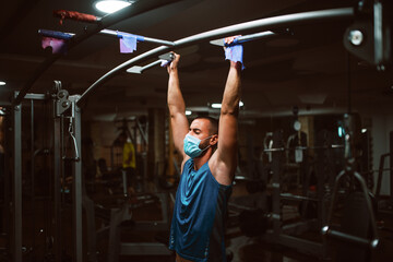 Muscular young fitness man with medical mask doing exercises on horizontal bar in a gym club. COVID 19 coronavirus protection