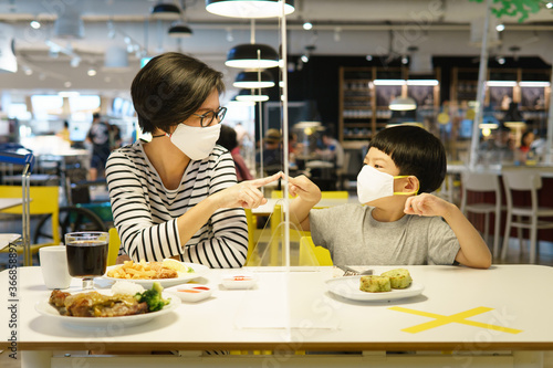 Stylish middle aged Asian mother and her child sit in food court reach out their index finger, touch on clear acrylic table barrier which separate them apart. New normal & Physical distancing concept.