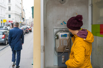 A young woman standing in front of an old payphone while calling someone. The street is right next to her with parked cars on the sidewalk.