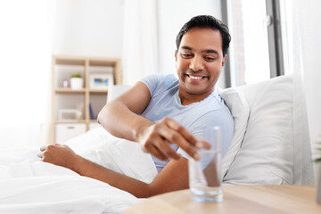 Poster - people, bedtime and rest concept - happy smiling indian man lying in bed at home and taking glass of water from table