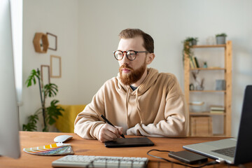 Wall Mural - Busy hipster cartoonist with beard sitting at wooden desk and using drawing tablet while making pictures