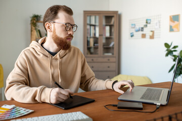 Wall Mural - Serious creative young man in hoodie working on design project using drawing pad in home office