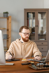 Wall Mural - Serious young bearded man in eyeglasses drawing design sketch on graphics tablet and editing it on laptop