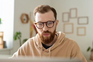 Wall Mural - Busy young bearded programmer in glasses working with laptop while analyzing pictures in home office