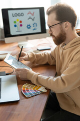 Wall Mural - Concentrated bearded man in hoodie sitting at wooden table and using tablet while working on brand design