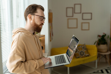 Wall Mural - Concentrated young bearded man in eyeglasses standing at window and analyzing interface sketch on laptop