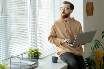 Wall Mural - Pensive young bearded man in hoodie sitting on window-sill and holding laptop while thinking of design
