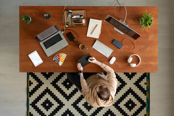 Wall Mural - Above view of interface designer in hoodie sitting at wooden desk full of devices and drawing sketch on digitizer tablet