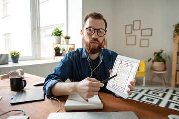 Wall Mural - Portrait of confident UI designer in eyeglasses pointing at tablet screen and presenting interface design while talking via video conferencing app