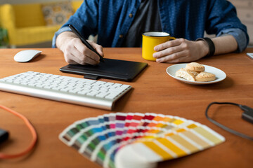 Wall Mural - Close-up of unrecognizable graphic designer sitting at table with mug and cookies on plate and working on visual product using digitizer tablet