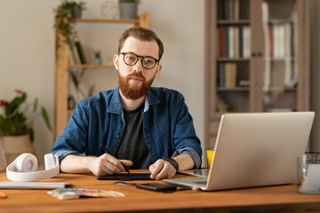 Wall Mural - Portrait of content handsome young male illustrator in eyeglasses sitting at table with drawing pad and laptop in home office