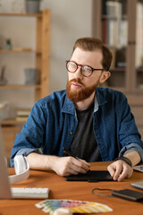Wall Mural - Hipster bearded man in eyeglasses sitting at table and using digitizer tablet while thinking of visual solution