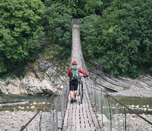 Man Walking With Cross Country Bike On Suspension Bridge Over River