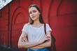 © BullRun - Portrait of attractive hipster girl in casual outfit standing on red wall background with crossed arms,beautiful traveler in white t-shirt with copy space for brand name or label posing outdoors