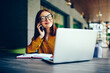 © BullRun - Portrait of positive female graphic designer in cool spectacles communicating via application on smartphone.Skilled redhead woman talking on cellular during remote work on laptop computer outdoors
