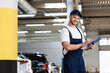 © LIGHTFIELD STUDIOS - happy mechanic in uniform and cap writing while holding clipboard and pen near cars