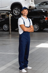 Wall Mural - cheerful mechanic in uniform and cap standing with crossed arms in service station